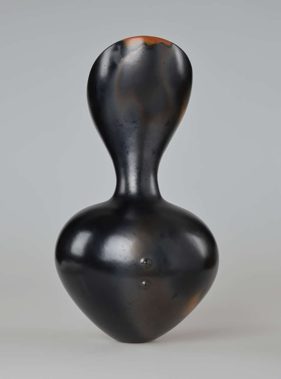 A black hourglass-shaped vessel with a smooth, glossy finish and small terracotta-colored patch on top. Two small raised bumps protrude from the center of the bottom half.