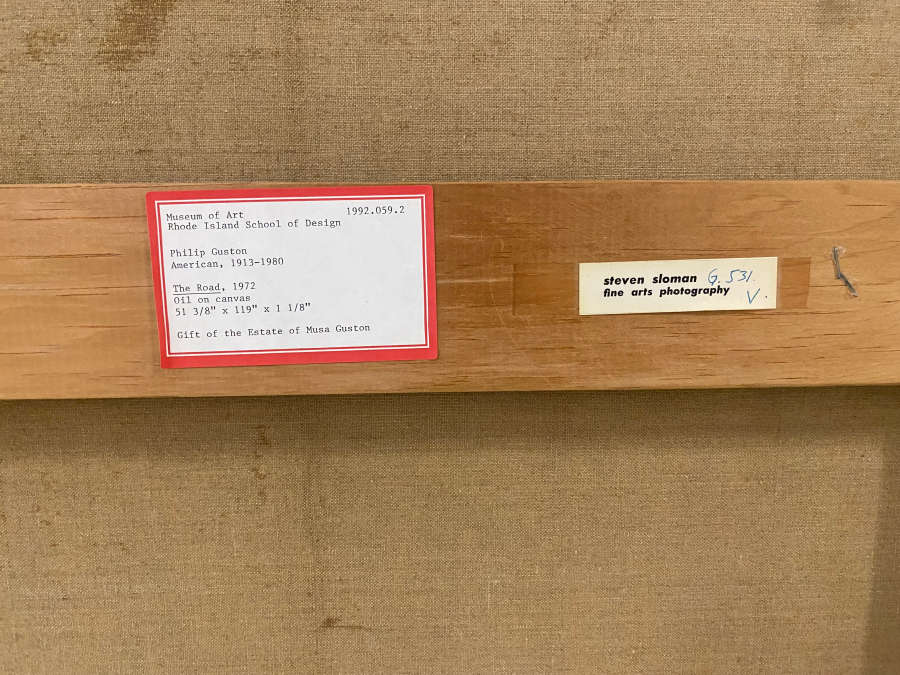 Back of a canvas with two labels on the wooden crossbar: a white museum label with text in a red border and a smaller cream photography label.
