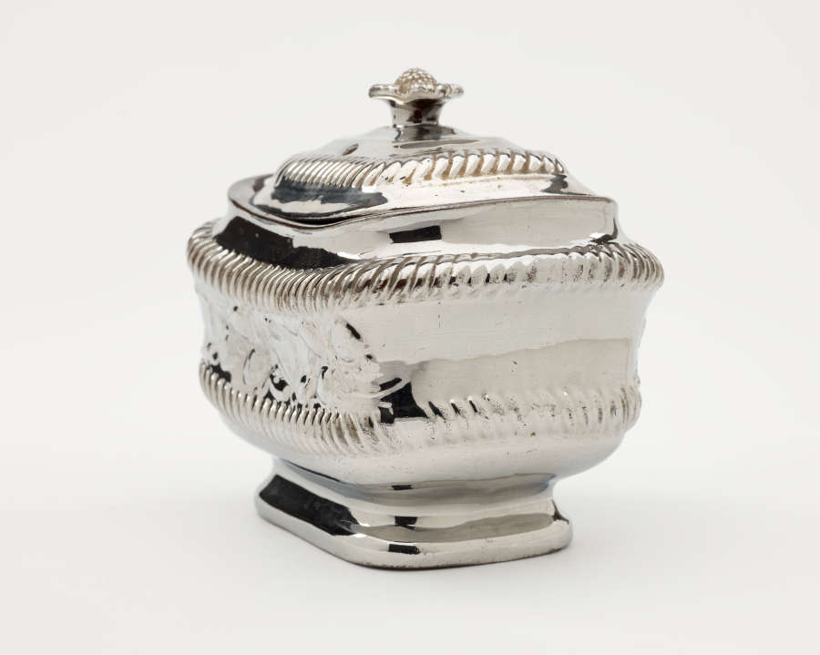 A silver sugar bowl with a decorative lid and a rounded square body with sculptural decorations.