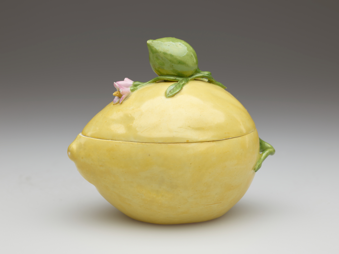 Lidded yellow box shaped like a lemon. Molded green leaves with a large bud and small pink flowers are applied to the top and wrap around the side.
