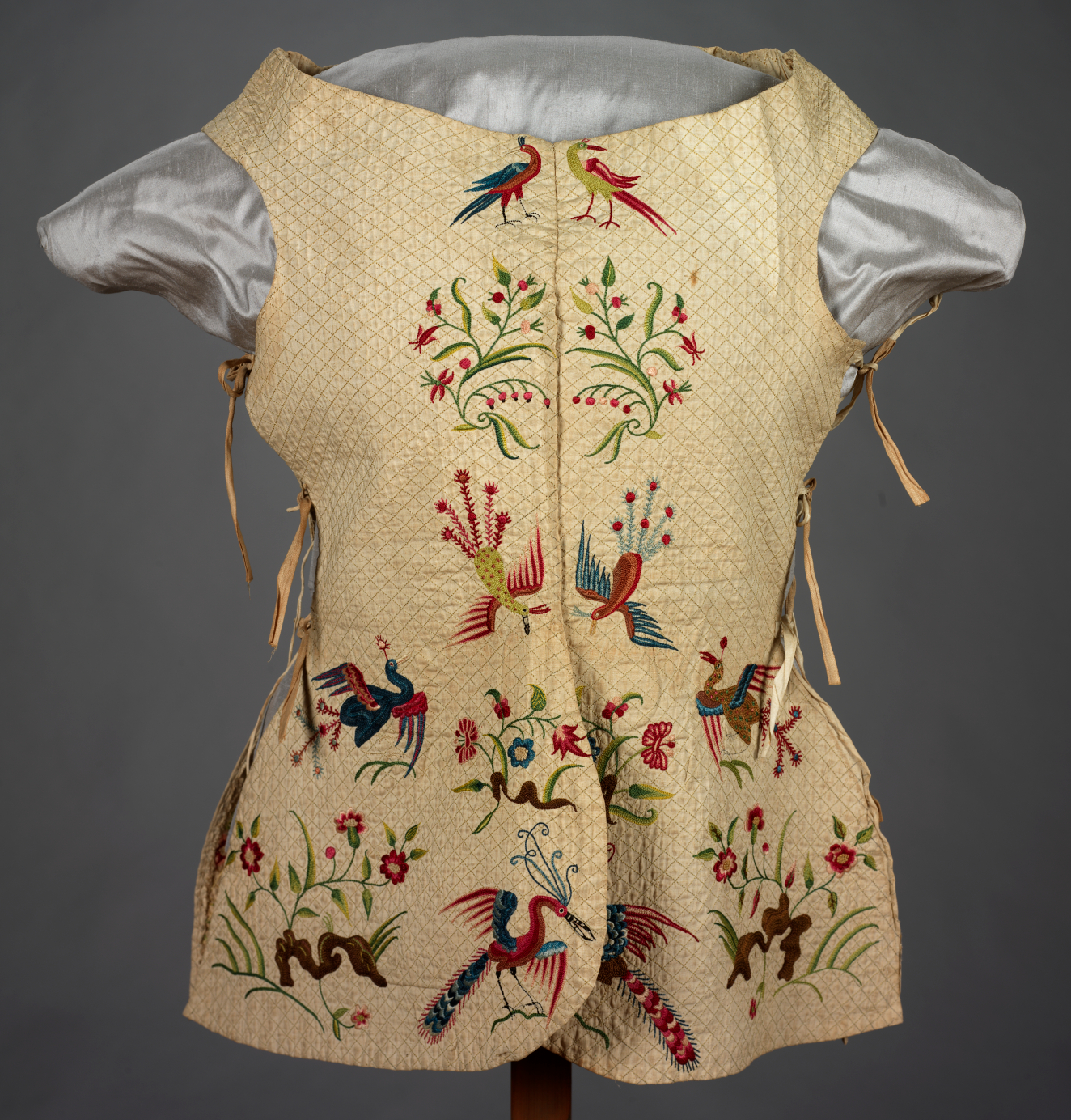 Front of a bodice with central seam decorated by a mirrored embroidered design depicting birds with plumage in varying combinations of yellow, red and blue besides delicate green foliage.