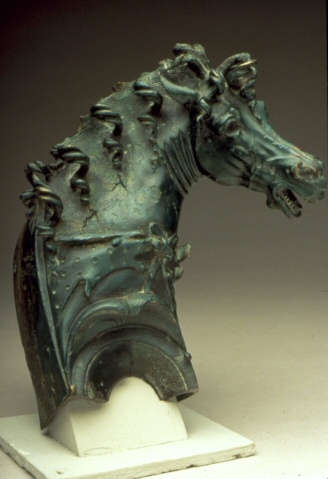 Greenish-gray metal ornament of a horse’s head fitted onto a white pedestal. Its head is turned, with curls of hair falling along the ridge of its neck and its saddle.
