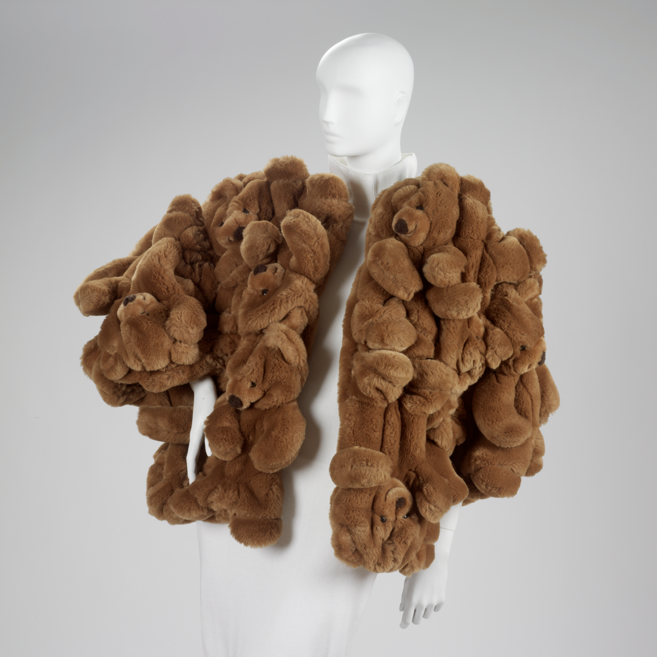 Bulky sculptural waist-length brown coat constructed entirely of densely packed plush teddy bear stuffed animal forms displayed on a white mannequin.