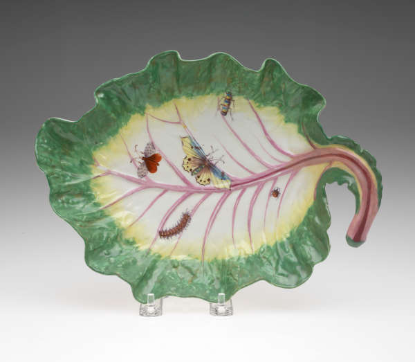 A leaf shaped dish in green, yellow, and white with a pink stem. Five insects depicted on plate: a lady bug, two butterflies, a caterpillar, and beetle.