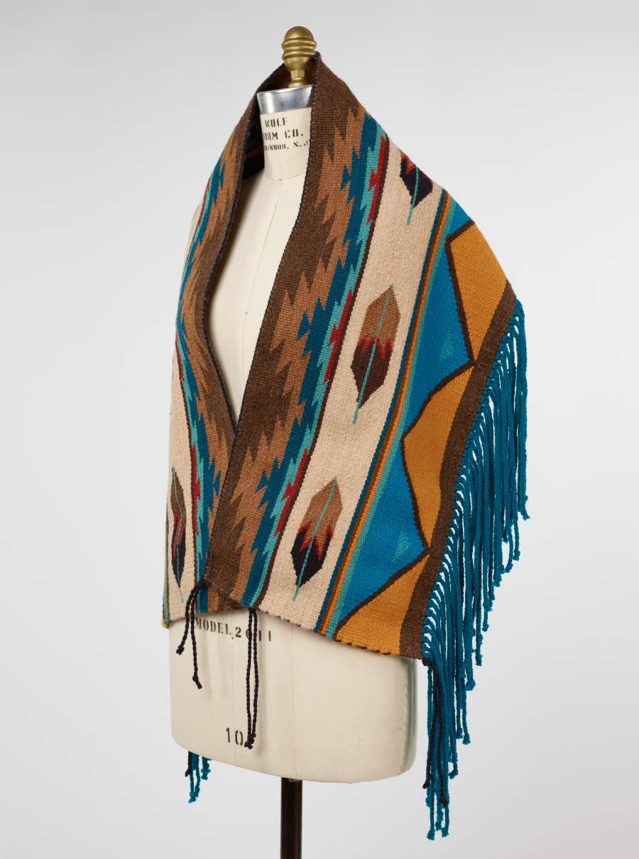 Side view of a mannequin wearing a multicolored woven fabric with fringe and tassels. 
The woven fabric contains brown feather motifs in between chevron designs and turquoise and yellow triangles. 