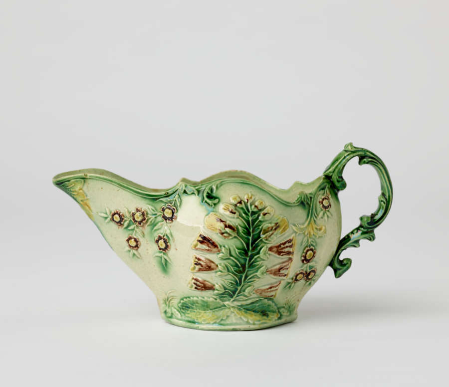  A short boat shaped pitcher green, cream, and brown in color with a sculptural handle and floral decorations.
