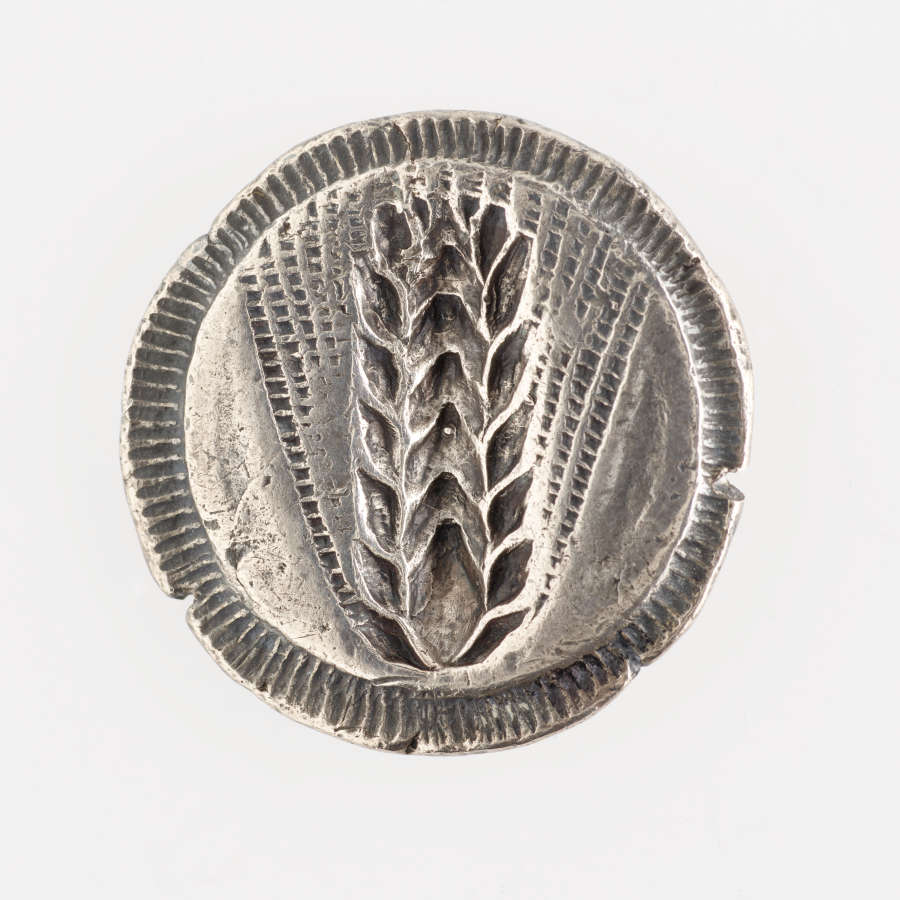 Round silver coin with irregular line-patterned edges, embossed with an image of a grain head of wheat with splayed awns.