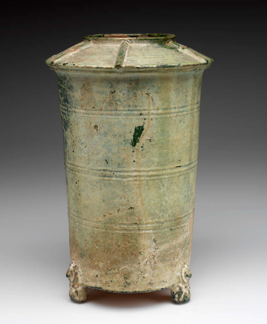 Side-view of a tall and narrow green-gray cylindrical terracotta urn with a narrow mouth and wide, upturned saucer-like neck, tapering into a narrow base, with one ornately carved foot visible.