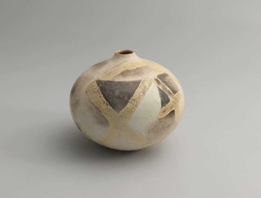 A spherical vessel with a narrow opening, patterned with muted beige, gray, and cream patches intersected by faded gold bands that create an abstract geometric pattern.