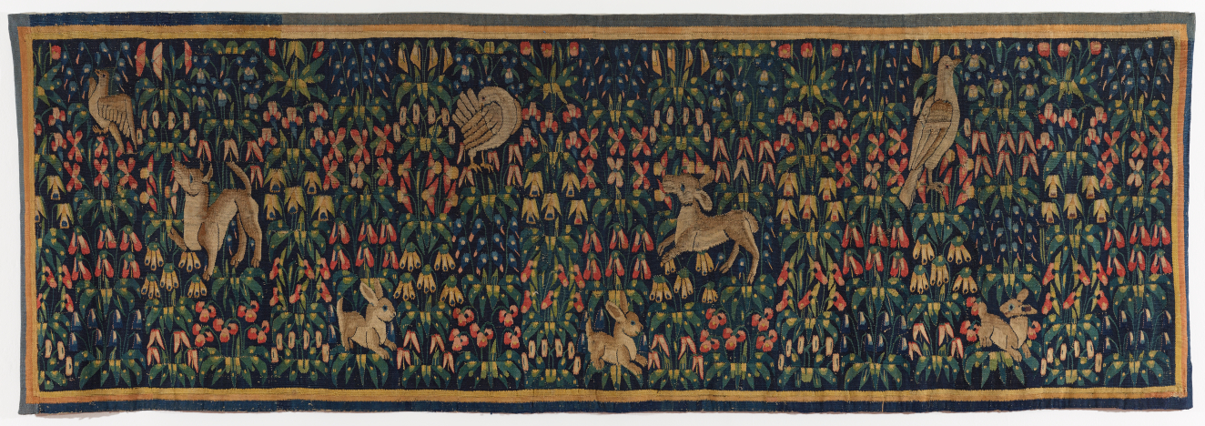Long, rectangular tapestry of a lush floral pattern of green, blue, red, and yellow. Golden animals including birds, deers, and rabbits are scattered throughout the dense foliage. 