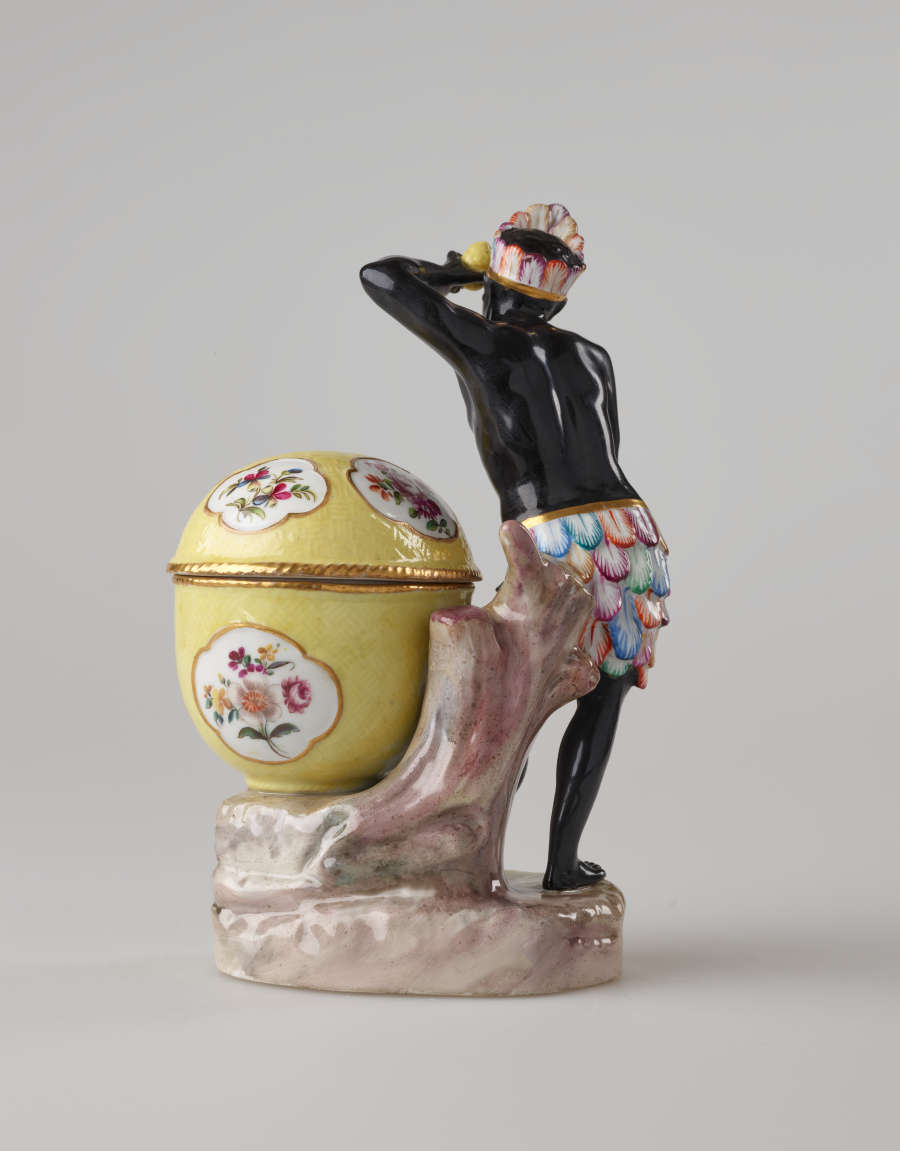 A sculptural figure with black skin, a colorful headdress and skirt. This figure is standing next to a bright yellow covered sugar basket with floral decorations.