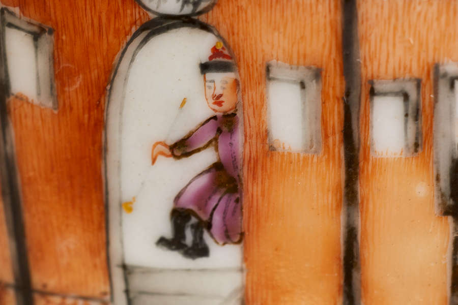 A close-up of a painted ceramic depicting a figure in a purple robe stepping through an arched doorway, set between an orange wall with small rectangular windows.