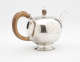 A bulbous silver teapot with a wooden finial and handle connected with decorative silver pieces. There are delicate engraved marks on the body and lid.