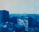 Monochrome blue elevated view of an early 20th century cityscape, featuring pitched roofs and chimneys. In the hazy distant horizon, several industrial structures can be seen.