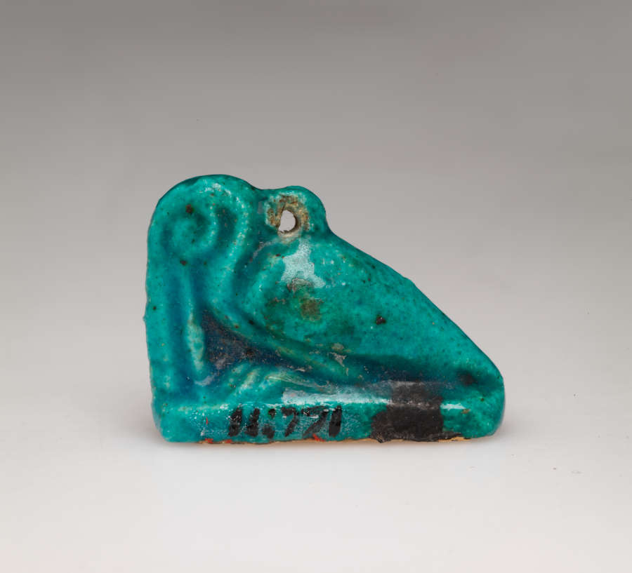 Turquoise amulet of a crouching pelican-like bird with a long beak pointed downwards, head tucked into its neck, where there is a small loop.  It is labeled in black ‘11.771’.