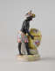 A sculptural figure with black skin, a colorful headdress and skirt. This figure is standing next to a bright yellow covered sugar basket with floral decorations.