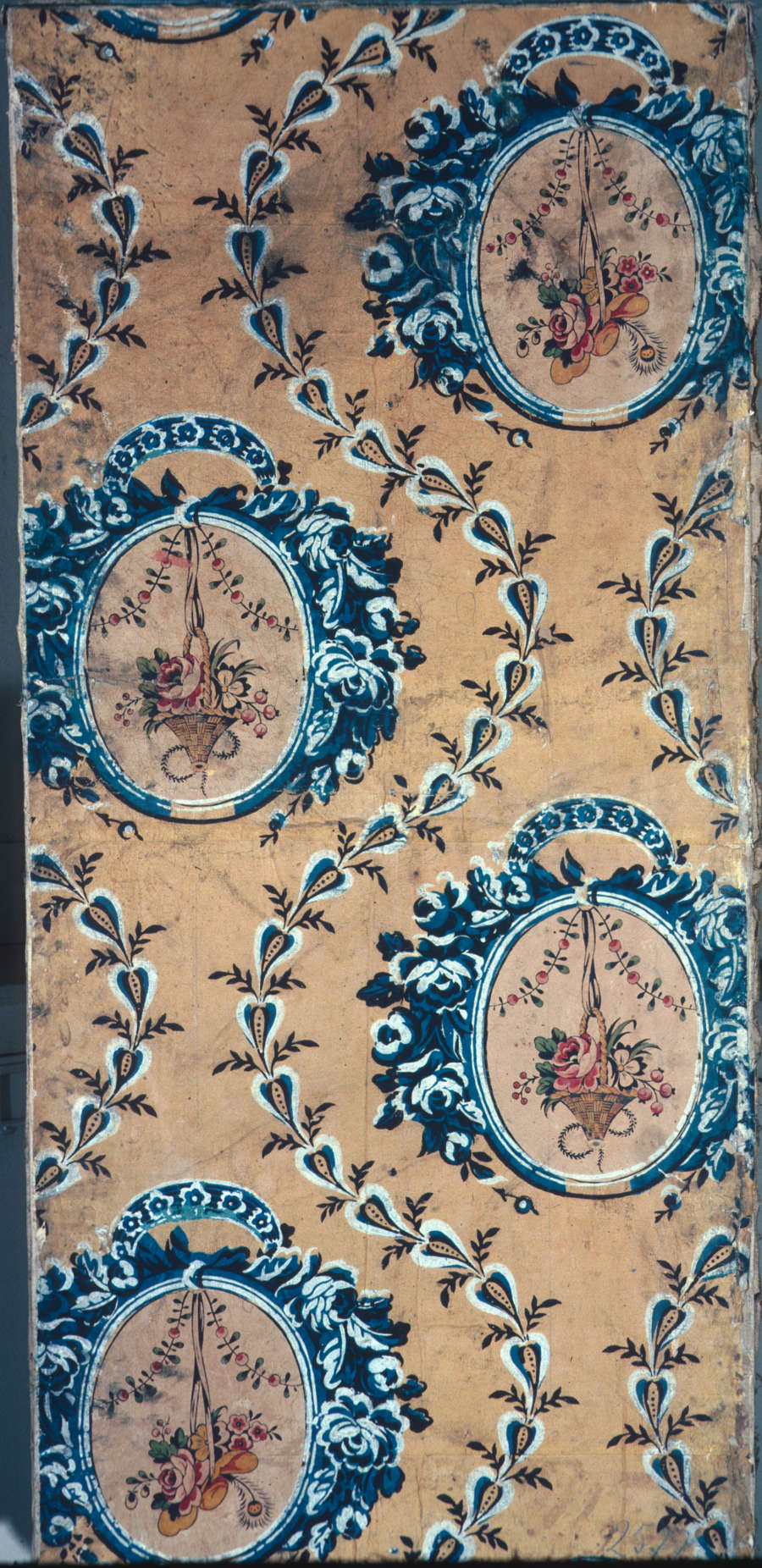 Aged wallpaper containing hanging floral bouquets within blue circular medallions against a pale orange background. Blue vine-like leaves curve around the medallions. 
