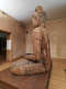 An angled view of a weathered wooden Buddha statue seated in meditation on a raised platform, in a warmly lit room.