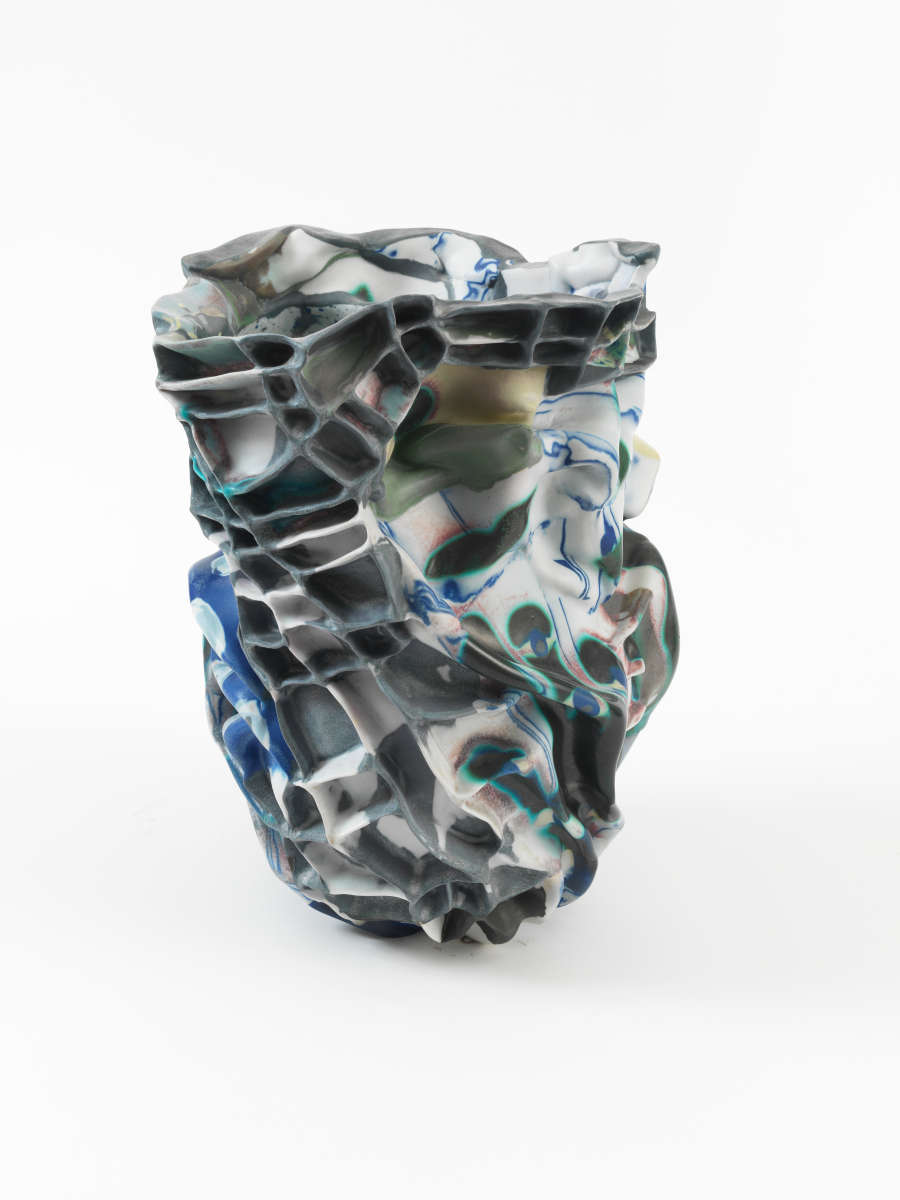 An abstract ceramic vessel constructed of mottled gray, white, and blue hollowed out grids that stretch and distort fluidly and irregularly, forming a jagged rim and a crumpled silhouette.