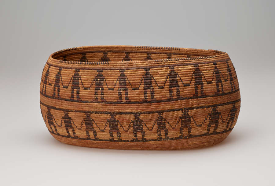 Side-view of an ochre-brown oval woven basket with irregular edges convex sides featuring two patterned stripes of dark-brown figures holding hands separated by horizontally running stripes.