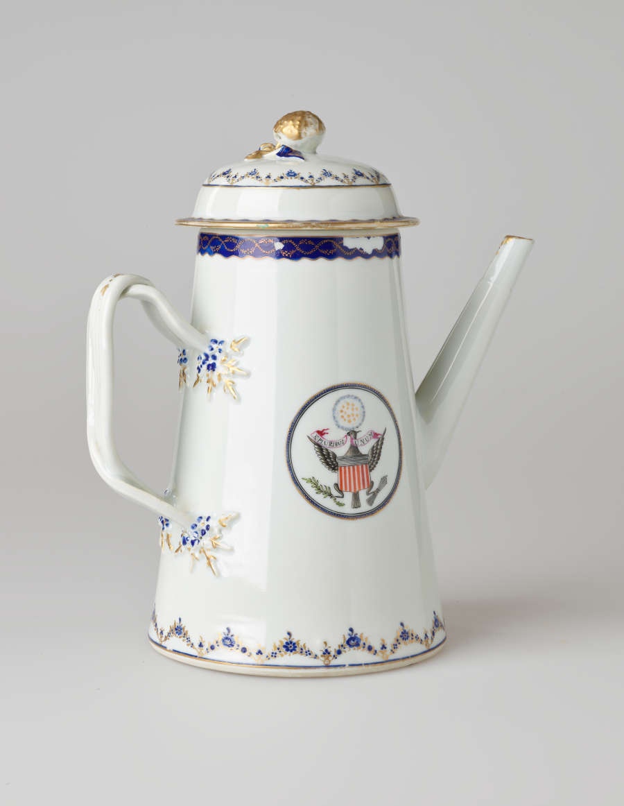 A white coffee pot with dark blue and gilded decorations, a straight spout, angular handle, and heraldry imagery in a circle on the body of the coffee pot.