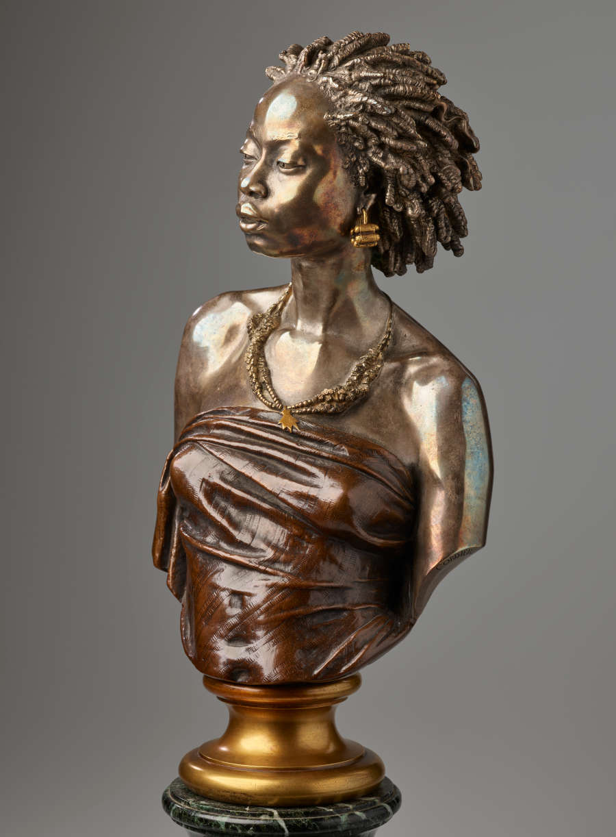 Bronze bust of a woman gazing downwards with hair in twists, gold earrings, beaded necklaces, adorning her collarbones above a dark strapless tightly-wrapped top mounted on a gold base.