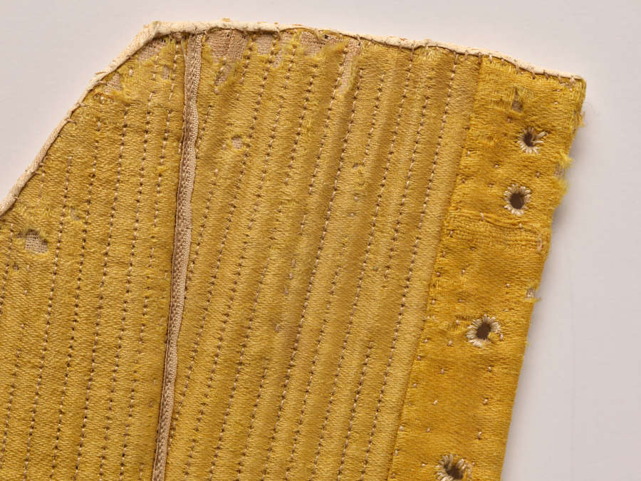Close-up photo of gold-colored fabric with many tiny stitches. The top is rounded and the right side has a row of small round holes.
