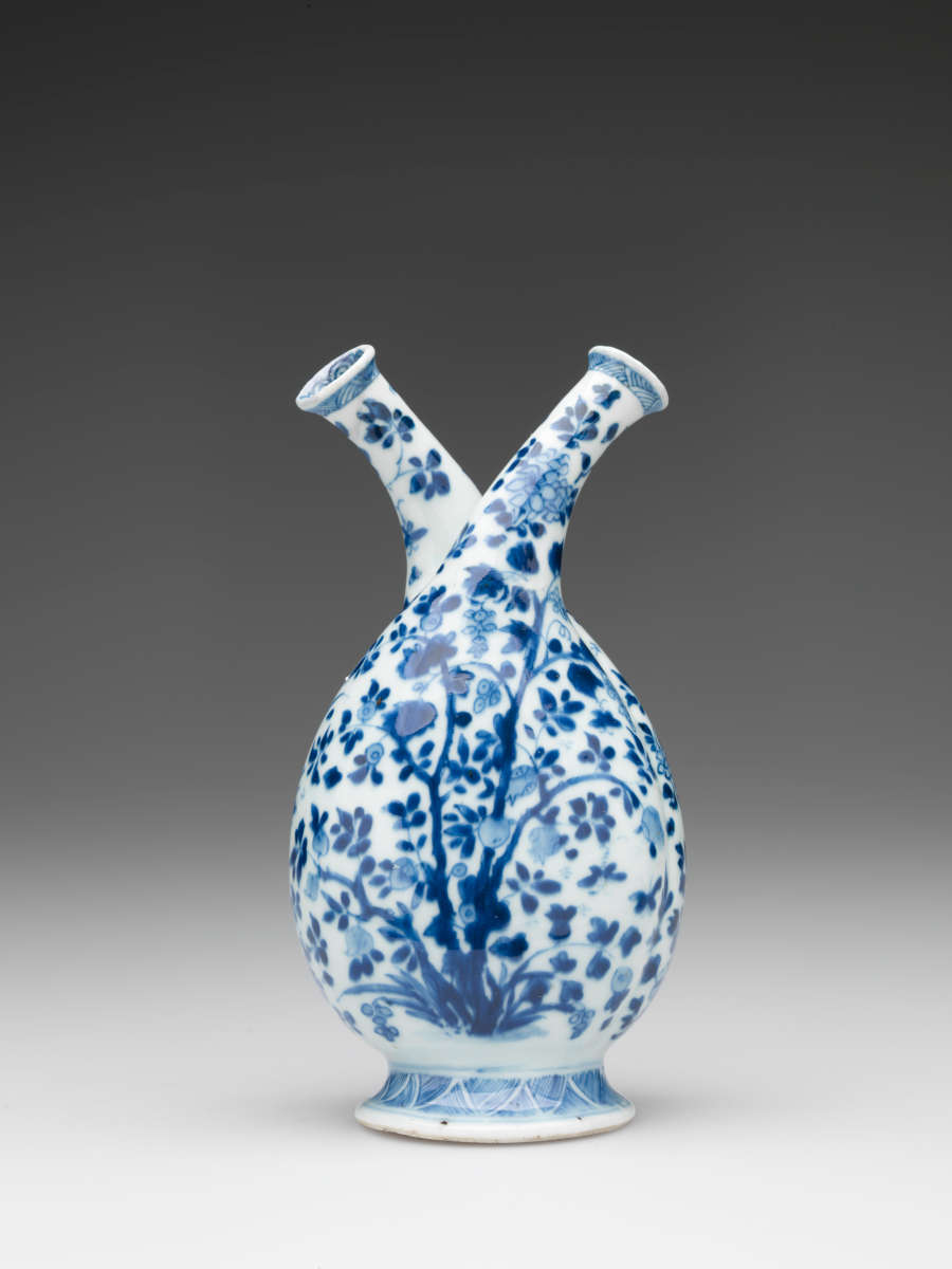 White bottle with two necks, patterned with dark blue leaves. Rounded at the bottom, the form tapers into two elegant spouts, which curve away from each other.