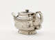 A silver teapot with a decorative handle, a rounded square body and spout with sculptural decorations.