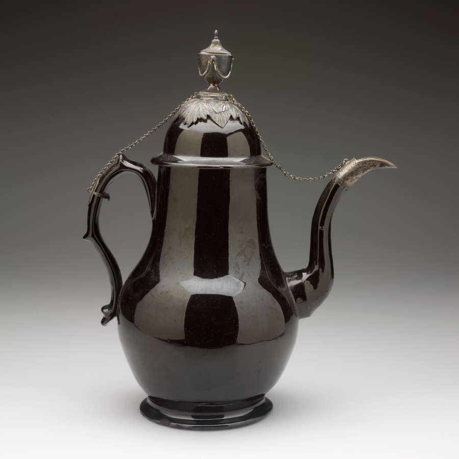 A black teapot with metal decorations on the spout, lid, and handle with a chain connecting them.