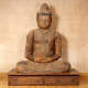 A weathered wooden Buddha statue sitting cross-legged on a wooden platform, with hands resting in the lap, with a serene expression and closed eyes. 