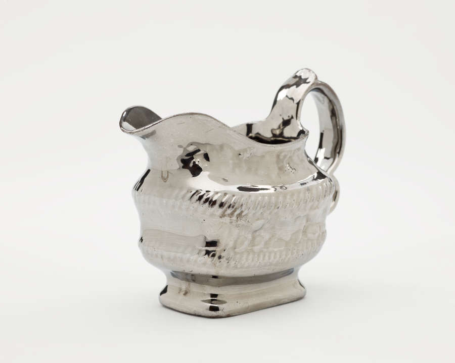  A silver creamer with a decorative handle and a rounded square body with sculptural decorations.
