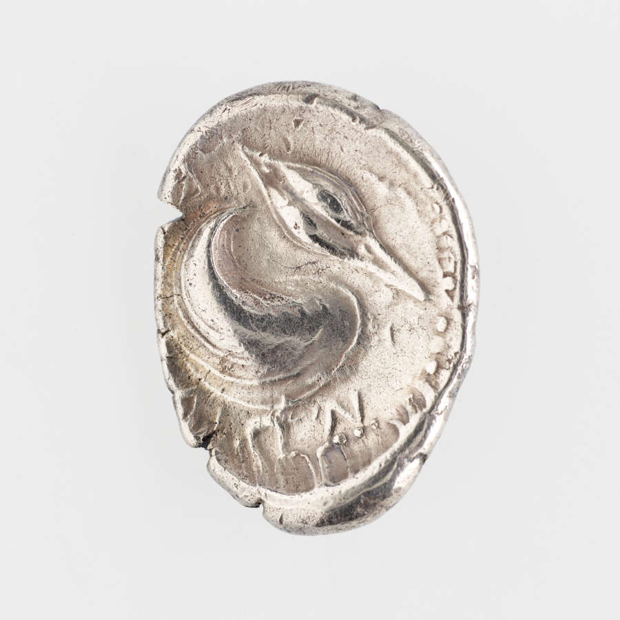 Other side of an ovular, rough-edged silver coin embossed with the profile of a swan-like bird, surrounded by Greek letters and a dotted border.