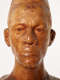 Close up front view of a glossy wooden bust depicting the head of a male figure with afrocentric facial features. He has a stern expression and an intense gaze.