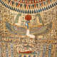 Painted coffin depicting a colorful winged human figure kneeling beneath a red sun with raised arms, bordered by hieroglyphs and decorative patterns.
