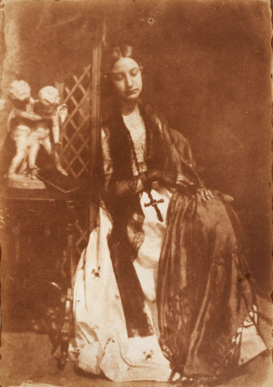 A warm brown portrait of a seated figure, eyes closed, with long hair holding a cross accompanied by a sculpture of two angels on the left.