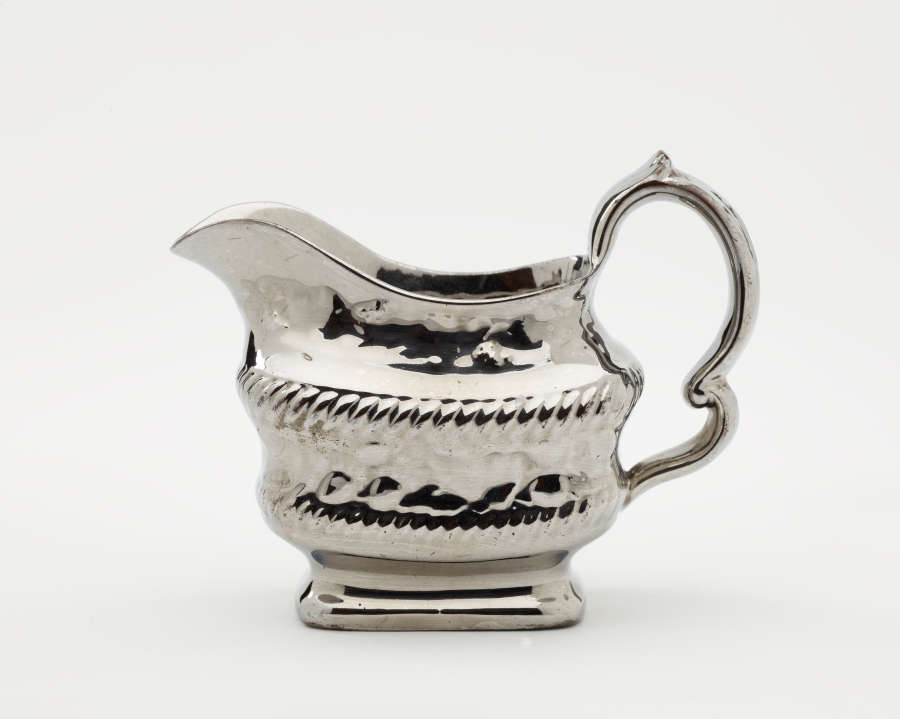  A silver creamer with a decorative handle and a rounded square body with sculptural decorations.
