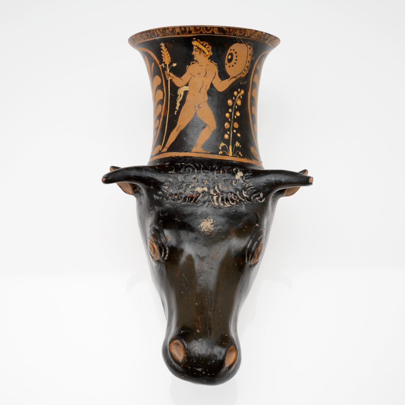 Black ceramic vessel shaped like a bull’s head with a flared cylindrical neck. The neck features an orange painted scene of a nude muscular figure holding a staff and shield. 
