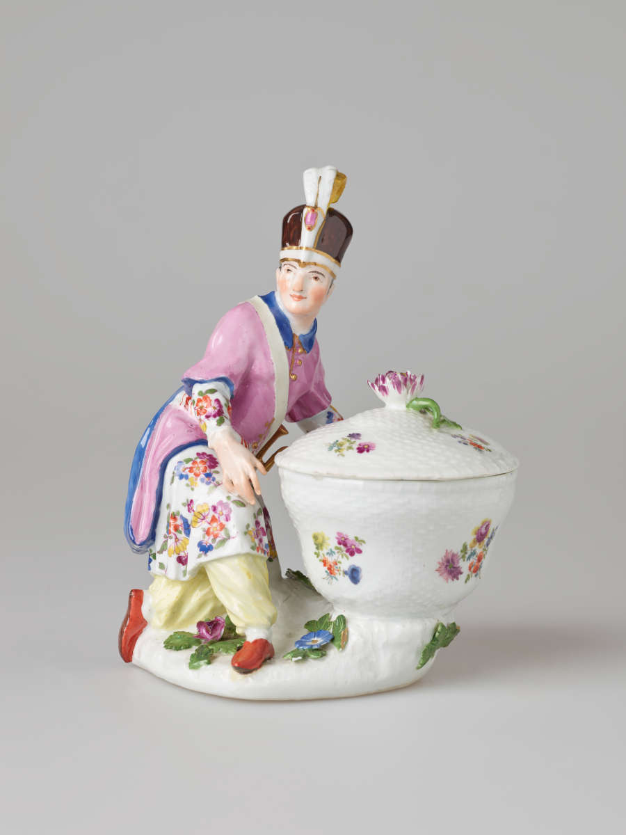 A sculptural figure kneeling next to a basket. Figure wears bright, elaborate clothing with a hat with a feather. There are floral decorations on the figure’s clothing and basket.