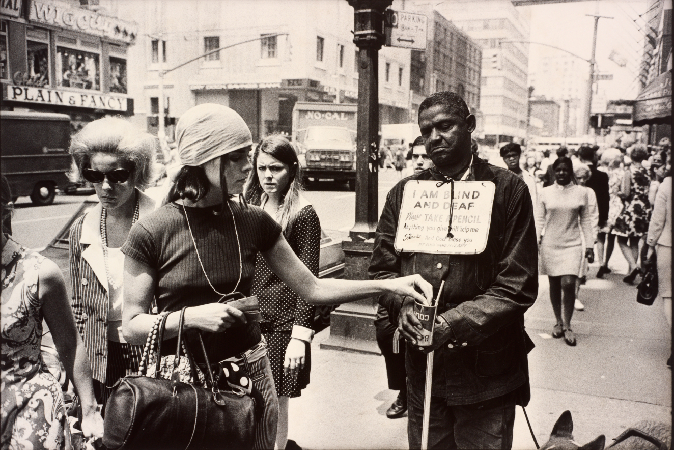 Black-and-white crowded street scene showing a dark-skinned person wearing a sign reading “I am blind and deaf” and a fair-skinned young person dropping a donation into his cup while another fair-skinned onlooker stares in disgust. 