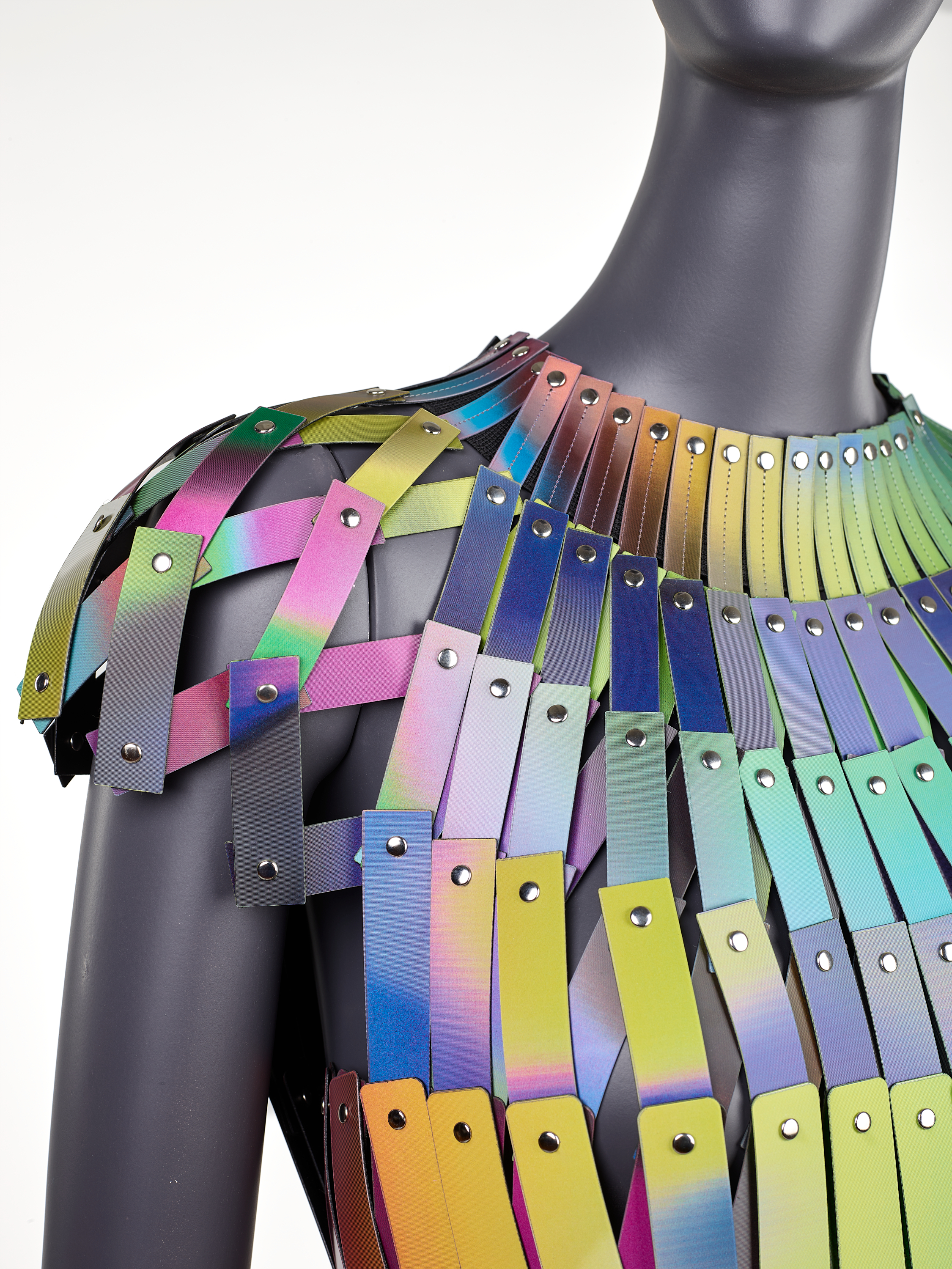 Close-up of a mannequin wearing a garment of overlapping holographic strips that shift in pink, yellow, and blue hues, each riveted in place to form a shimmering scale-like pattern. 