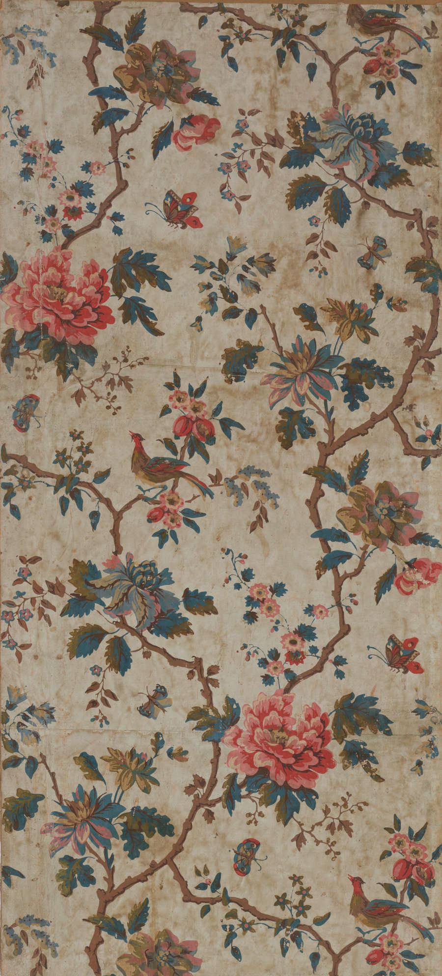 Tan, aged wallpaper with repeating motifs of multicolored flowers and leaves on brown jagged branches. Tiny, colorful butterflies and birds on branches repeat throughout. 