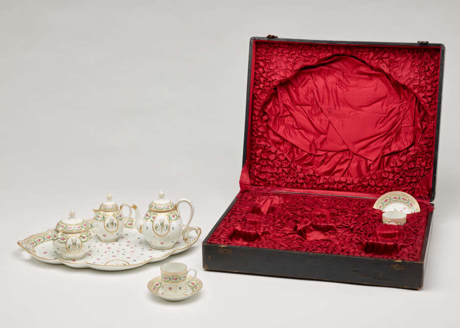 A white, pink, green, and gilded tea set with floral decorations and consists of four vessels and a tray. The traveling case with a black exterior and red fabric interior.