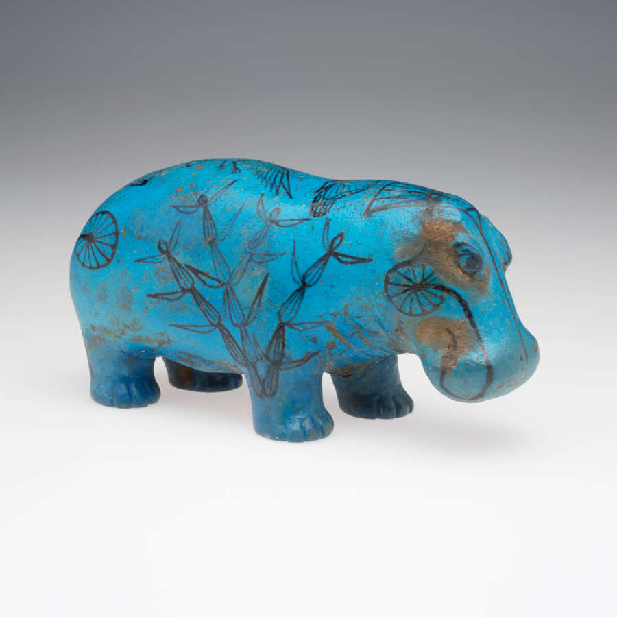 Quarter view of a blue sculpture of a standing hippopotamus with black outlines of birds, insects, plants and waves on its glossy surface. It stands against a gray background.