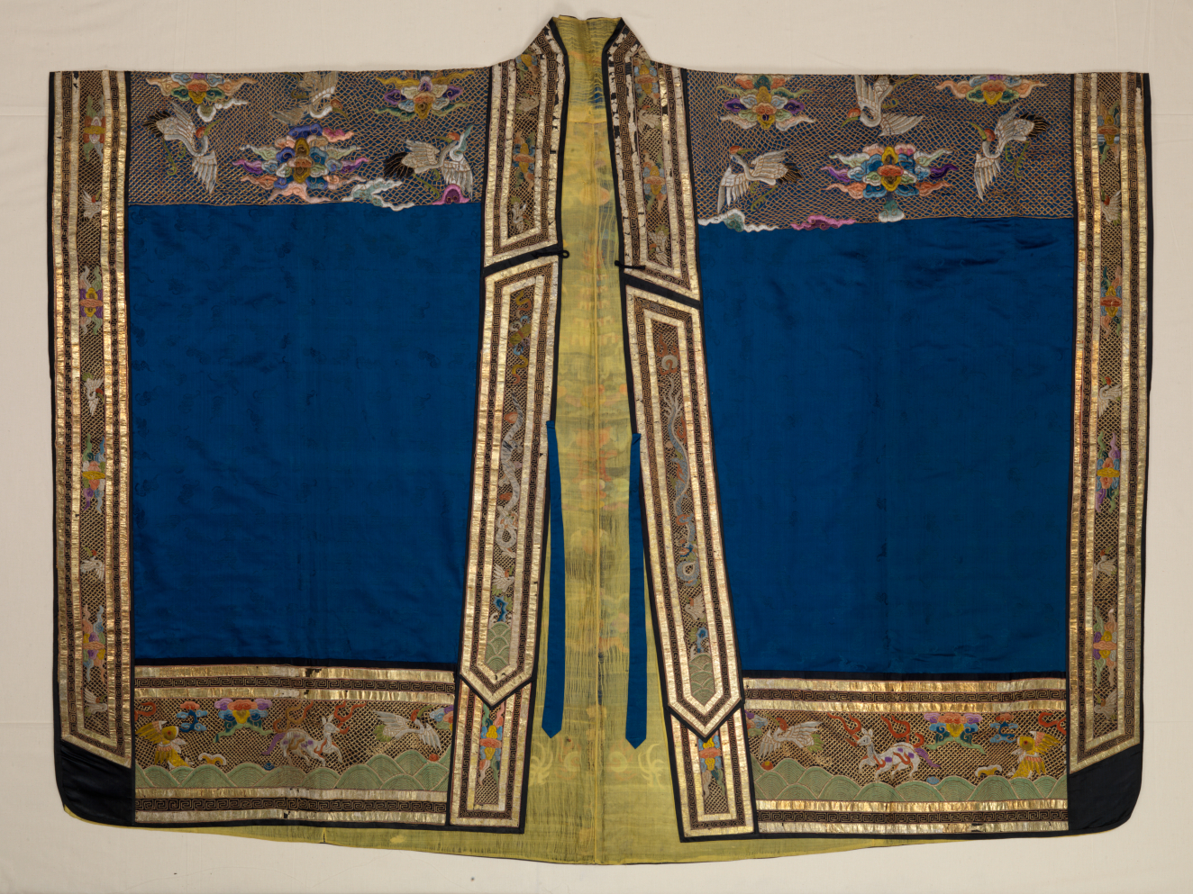 A richly decorated robe with broad sleeves, with intricate embroidery of cranes in flight, colorful blossoms, and geometric patterns against a shimmering gold border, framing a blue center. 