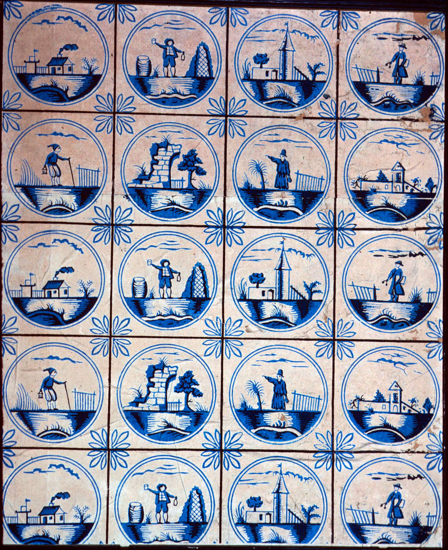 Tan wallpaper with eight, blue circular-framed repeating motifs of buildings and figures working, all on a bridge, arranged in a 5 by 4 grid. outlined flowers line the grid’s intersections.