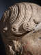 Close-up of a carved stone head focusing on curled locks of hair that coil at the end. The deep grooves are illuminated to highlight the craftsmanship. 