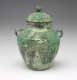 A closed jug container with a round bulbous body, short neck, round lid, and two handles. The surface features patches of different greens, and carvings of geometric patterns and characters.