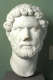 Marble bust from the neck up of a realistically carved bearded man with loose curly hair and furrowed brows resting on a pedestal against a gray backdrop.