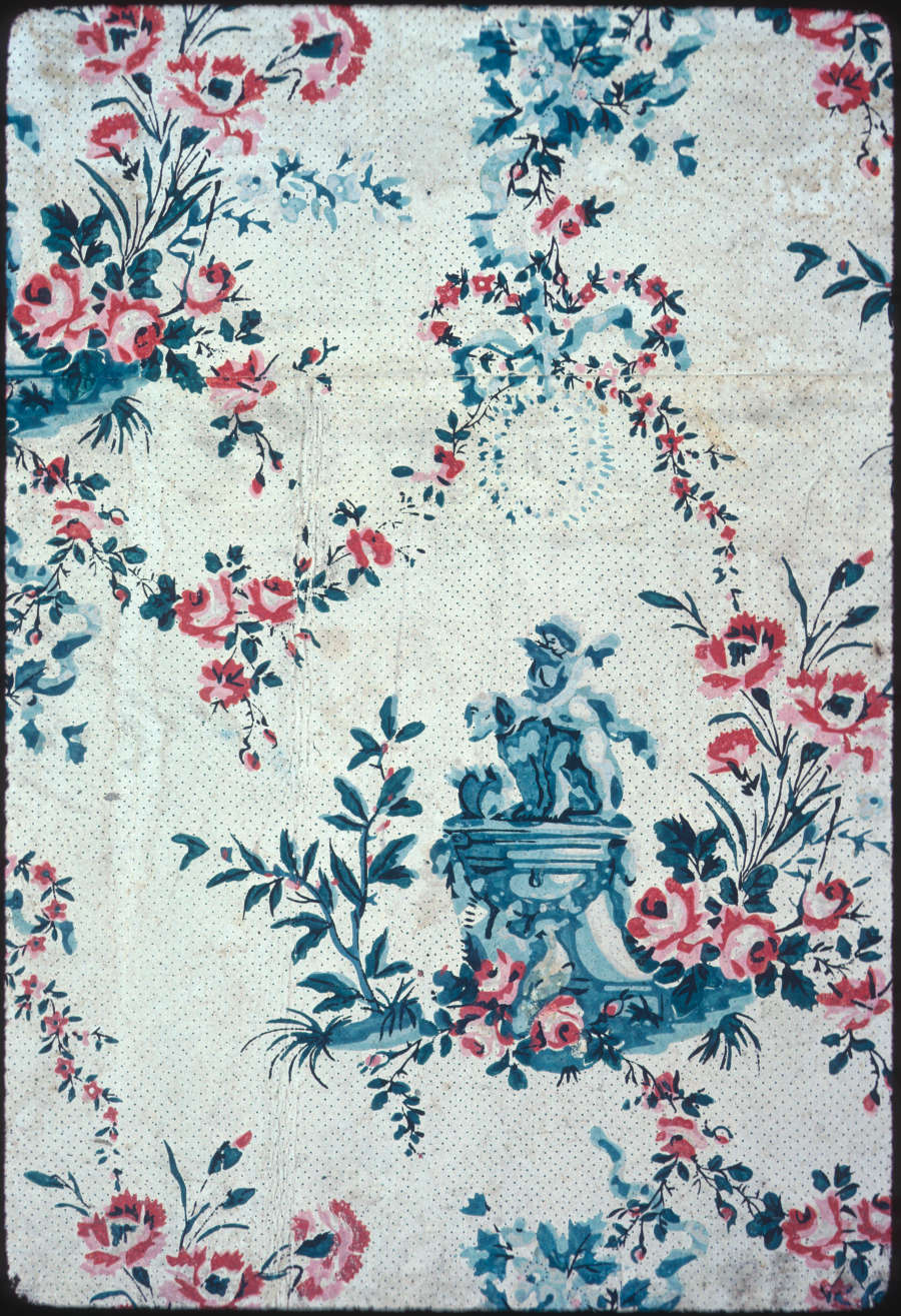 Aged, polka-dotted cream wallpaper with repeating light gray winged baby statues standing on a pedestal. Stemmed salmon pink flowers and floral garlands, which vary in size, surround the pedestal.